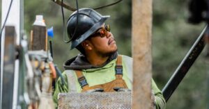 Construction worker wearing a hard hat, safety glasses, and green overalls, looking up while handling a perforated metal panel at a job site.